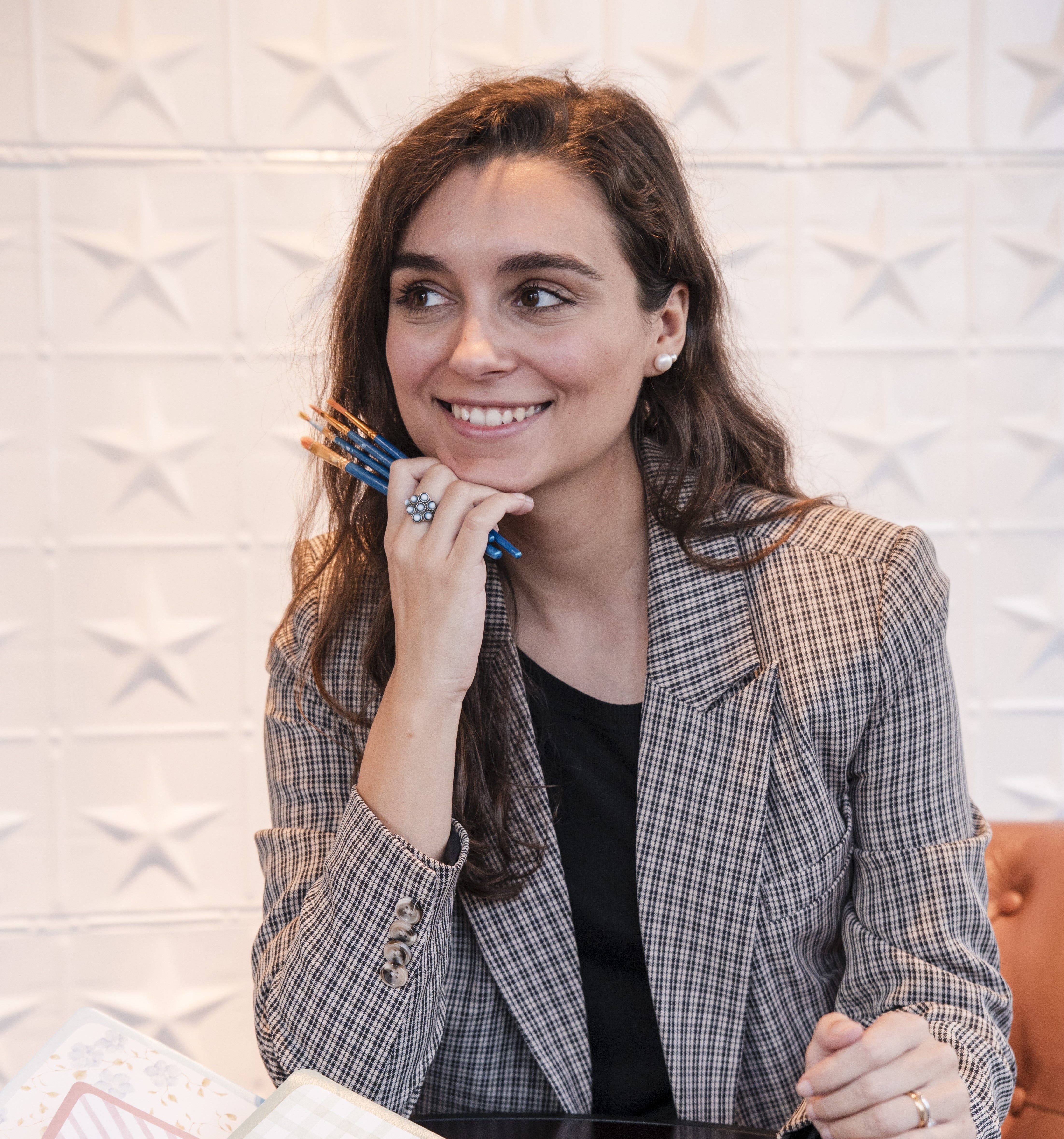 Woman in a checkered blazer holding painting brushes with a textured white wall background