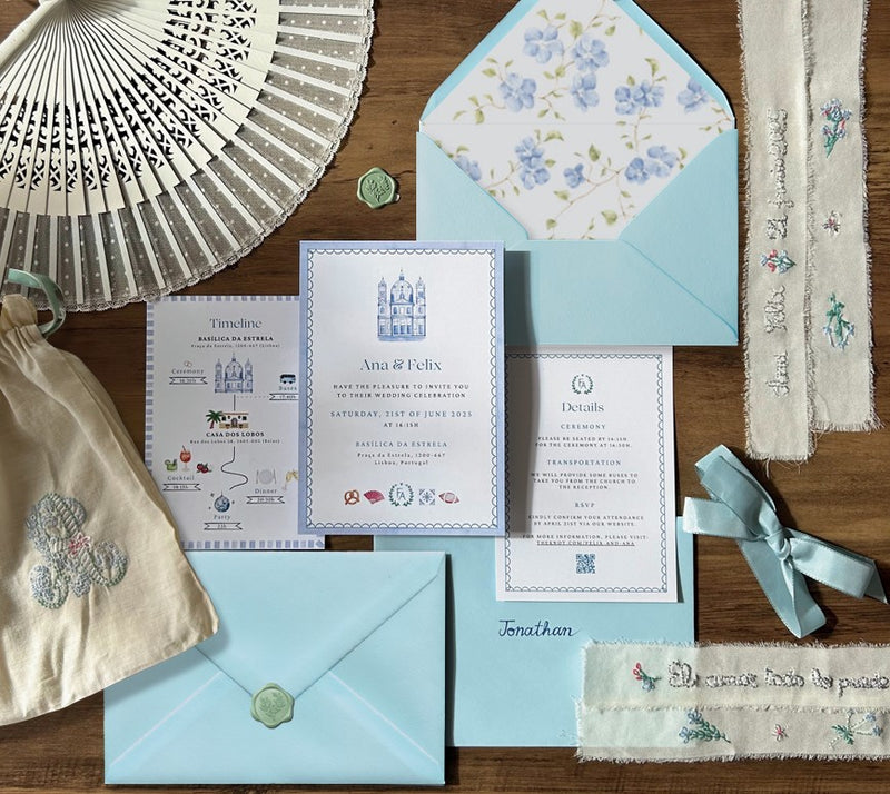 Wedding invitation set with blue envelopes, decorative paper, and a fan on a wooden surface.