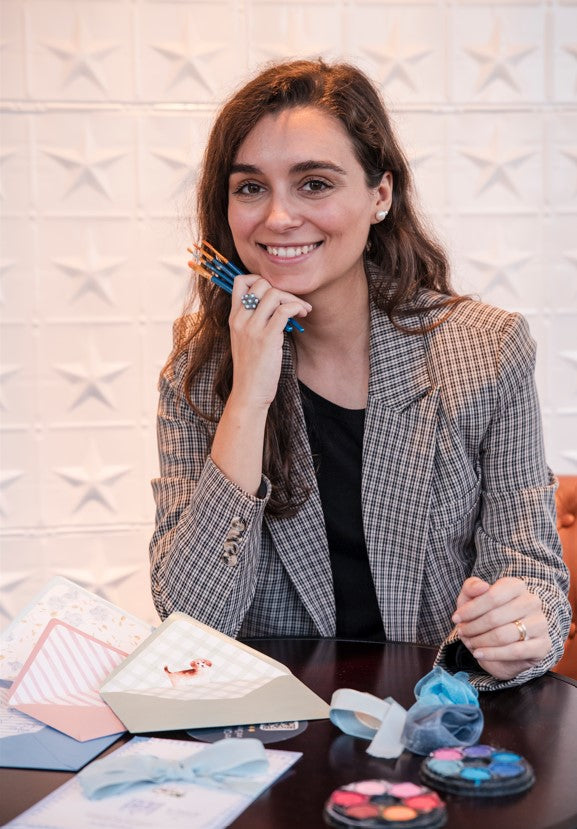 Woman smiling sitting at a table, with painting brushes, invitations and watercolors.