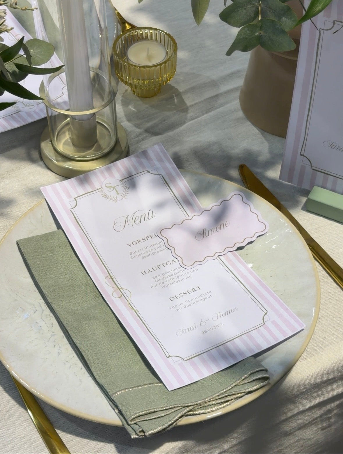 Table setting with a pink and green menu and name card on a table, featuring greenery and gold accents.