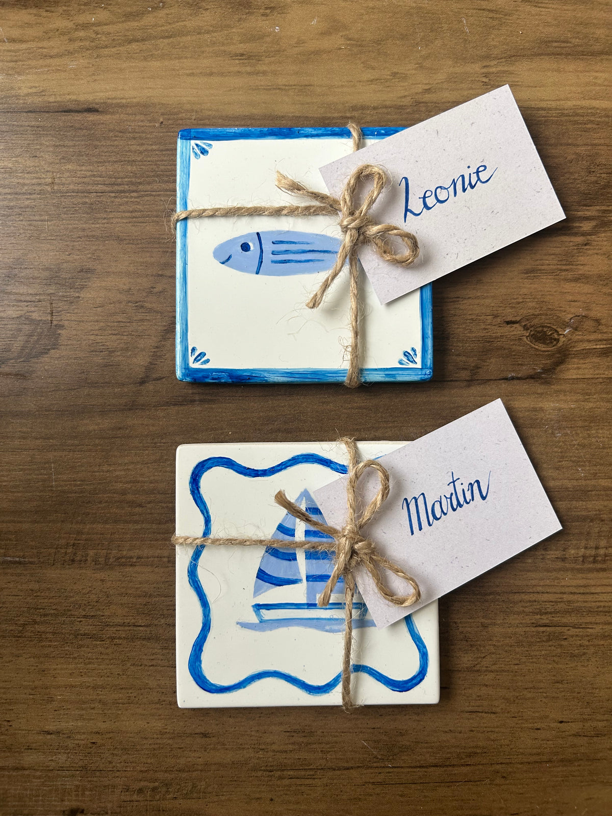 Two handpainted tiles with name cards tied with string on a wooden surface